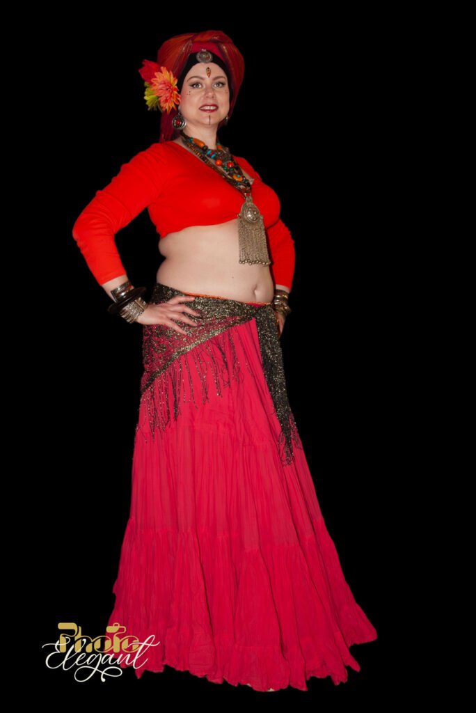 Gypsy woman in a traditional red dress and headscarf, wearing silver fashion jewelry, standing against a black studio backdrop.