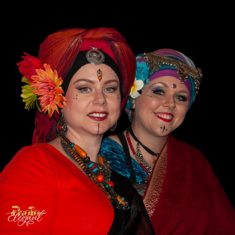 Two young gypsy women in red traditional dresses with floral headscarves and silver jewelry on a black background