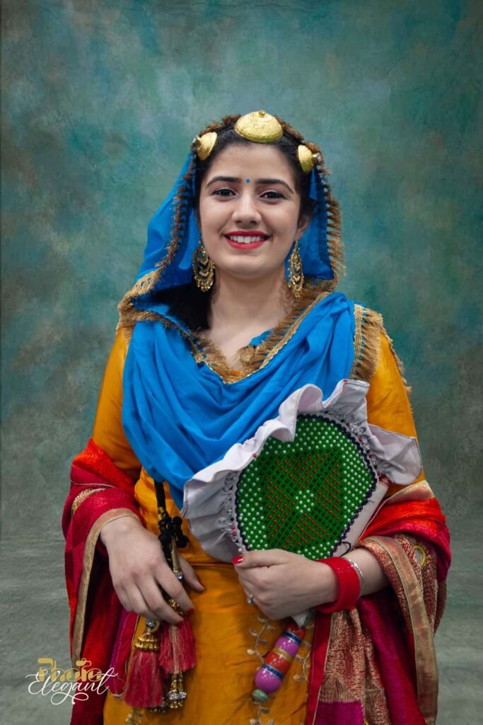 Smiling Indian girl in colorful traditional dress holding a hand-held wooden fan, standing against a turquoise backdrop in a studio.