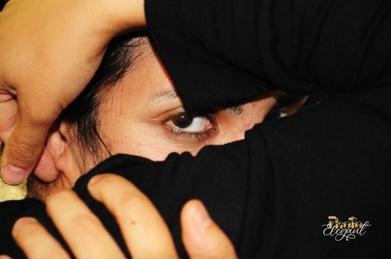 A beautiful young woman hides her face with her arm, revealing only her striking eyes and intense gaze.