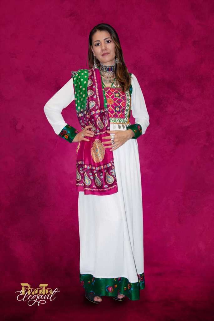 Elegant Afghan woman in vibrant traditional dress with silver necklace and earrings, seated on a sofa against a red backdrop.